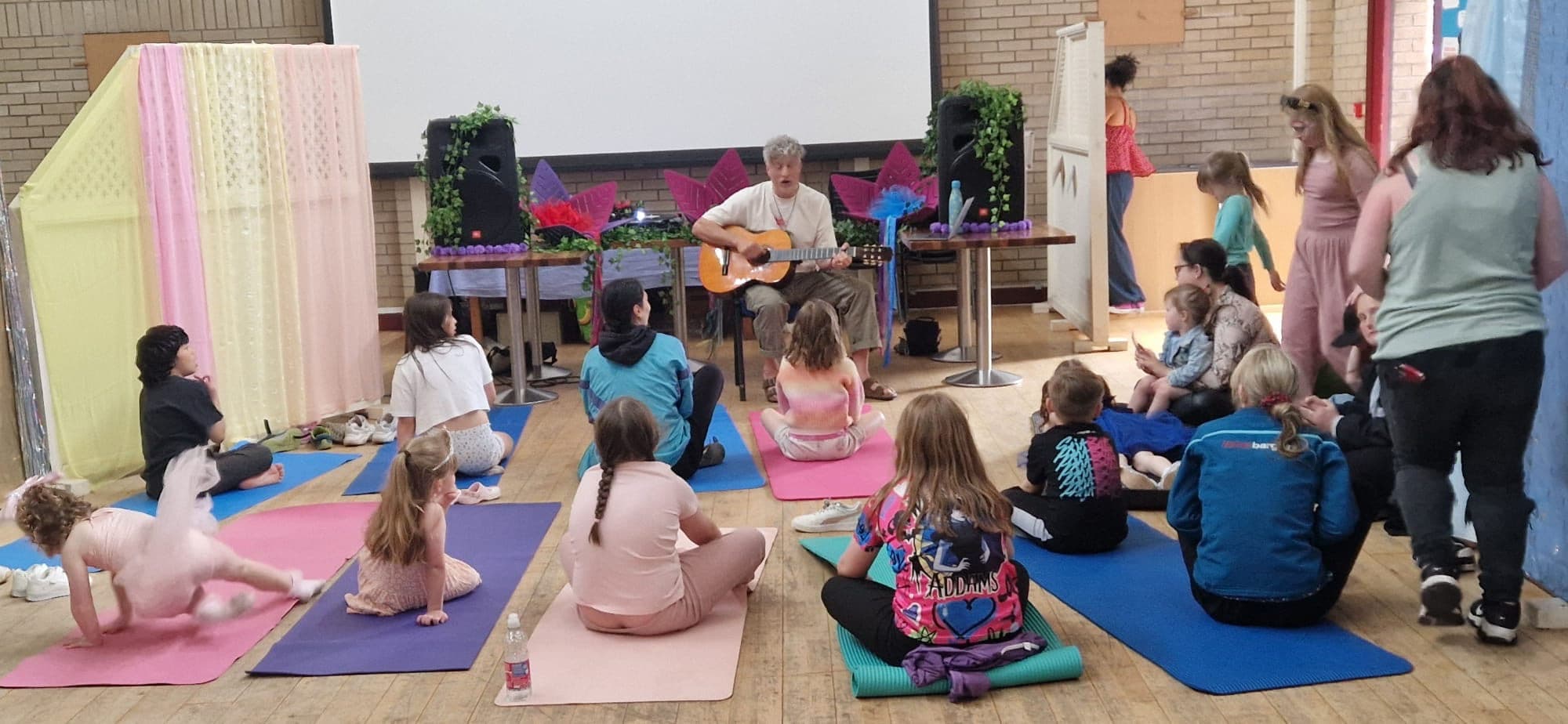 Children and grown-ups gathered in a circle around a guitar, singing together.