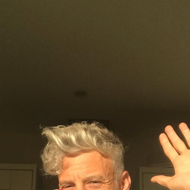 A grey-haired man grinning up at the camera in a low-angle selfie, one hand raised mid-wave in warm indoor light